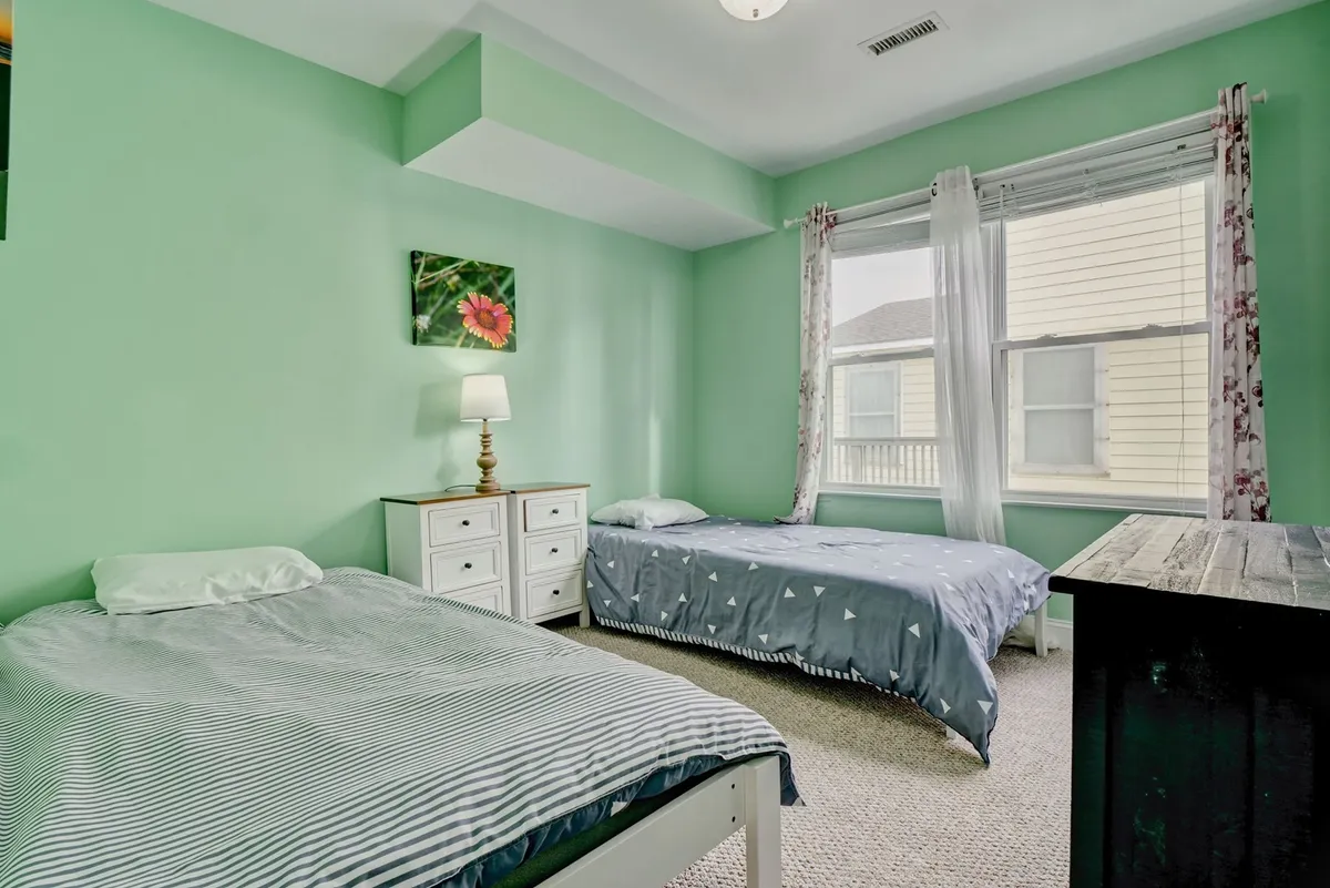A welcoming guest bedroom with green walls, two beds featuring patterned bedding, a bedside lamp, and floral artwork. The layout feels comfortable and flexible, suitable for families or friends traveling together.