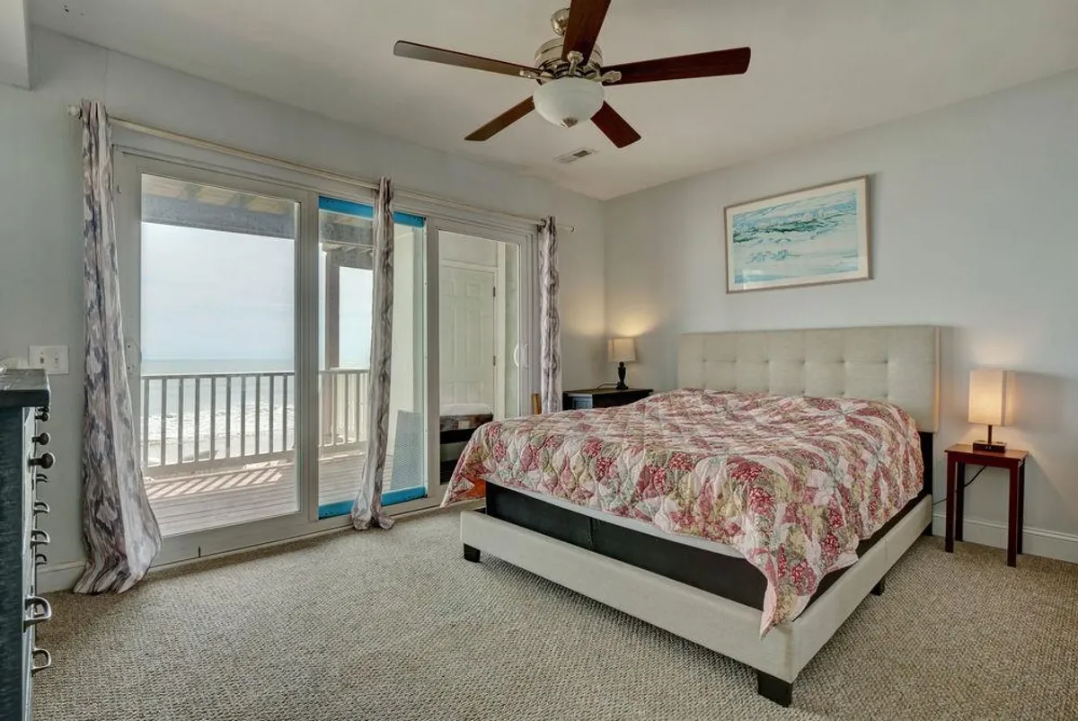 A tranquil bedroom featuring a floral quilted bed, large windows with an ocean view, a ceiling fan, and a side table with a lamp. The space is bathed in soft light, creating a cozy and restful environment ideal for unwinding after a day at the beach.