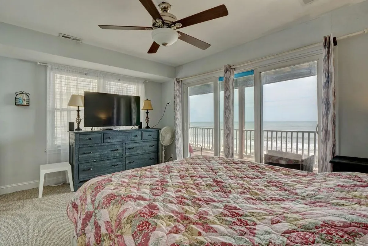 A sunlit master bedroom with a cozy quilted bed, large windows offering an expansive ocean view, and a dresser with a television. The calm mood is enhanced by soothing colors and natural light, emphasizing serenity and comfort.