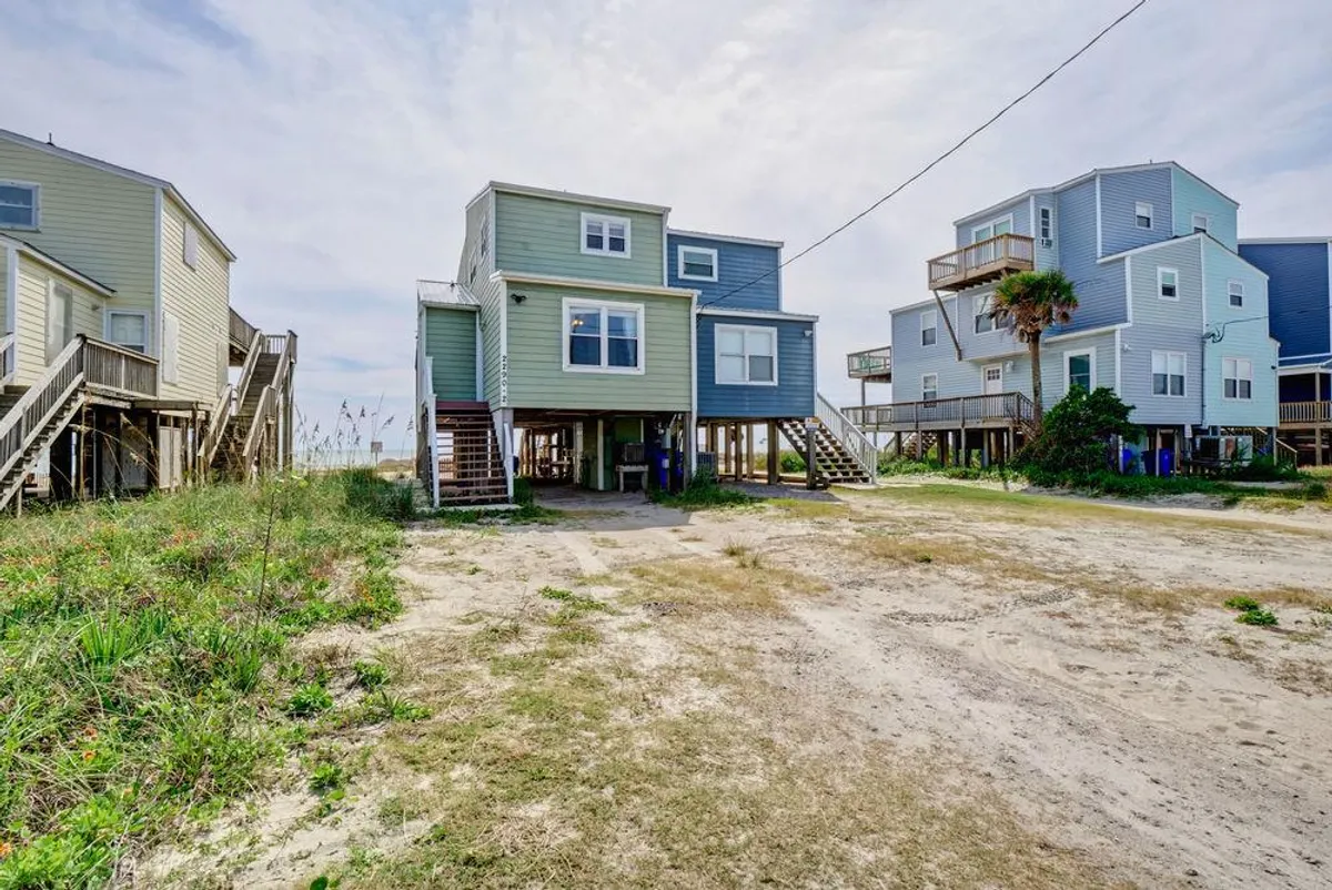 An exterior shot of colorful beach houses with stilt architecture, surrounded by lush greenery. The image captures ample driveway space and carports beneath the homes, hinting at parking convenience and a relaxed, welcoming atmosphere.