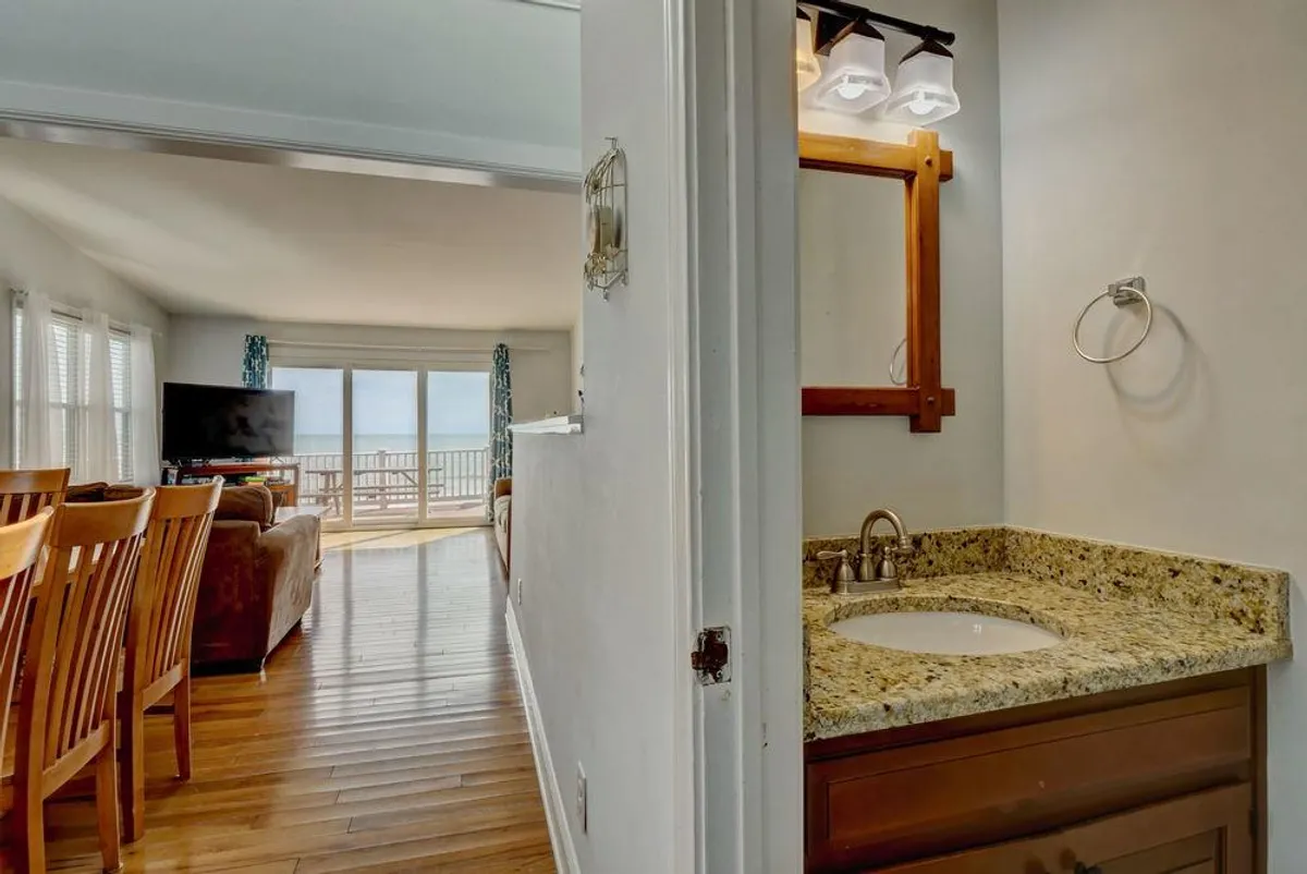 This image presents a welcoming, sunlit open-concept living area with large windows offering ocean views. The scene includes a cozy living space with a couch and television, a dining area featuring wooden chairs, and the hint of a bathroom vanity, all bathed in natural light. The overall mood is relaxed and sociable, ideal for family gatherings or casual relaxation.