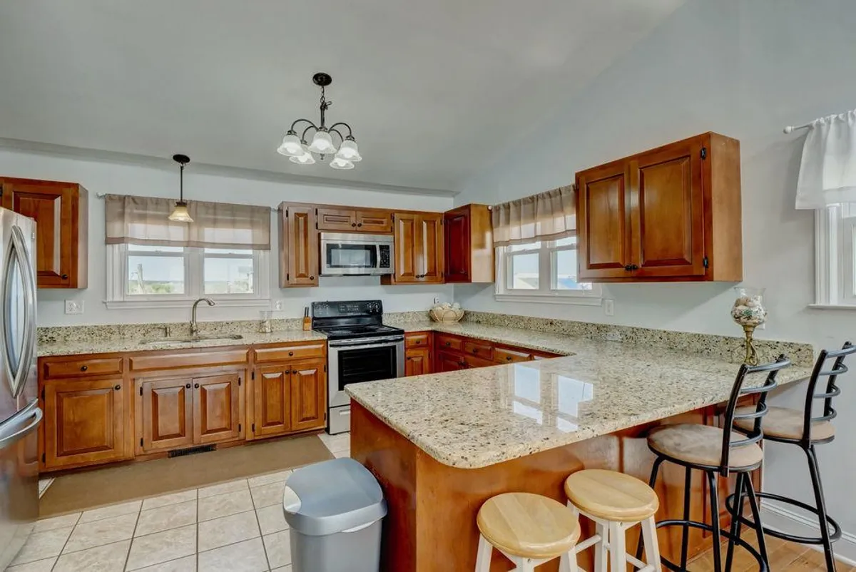 This image showcases a spacious, open kitchen with granite countertops, wooden cabinetry, stainless steel appliances, and bar stools lined up along the island. The layout encourages communal cooking, casual dining, and group gatherings, all in a modern and inviting atmosphere.