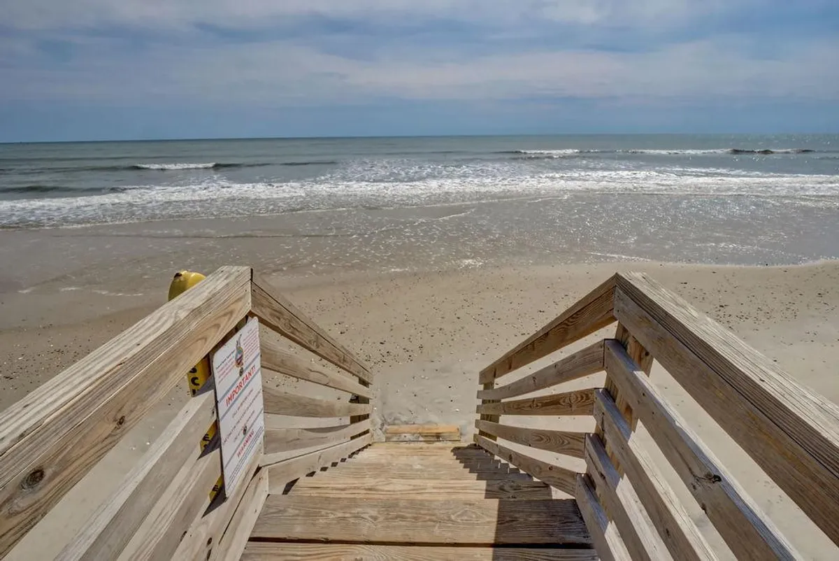 Wooden steps descend directly onto sandy beach, with ocean waves and an expansive sky in view. The scene captures the transition from the property’s deck area to the public beach, embodying effortless beach access. The open, sunlit setting suggests easy movement between home and shoreline.