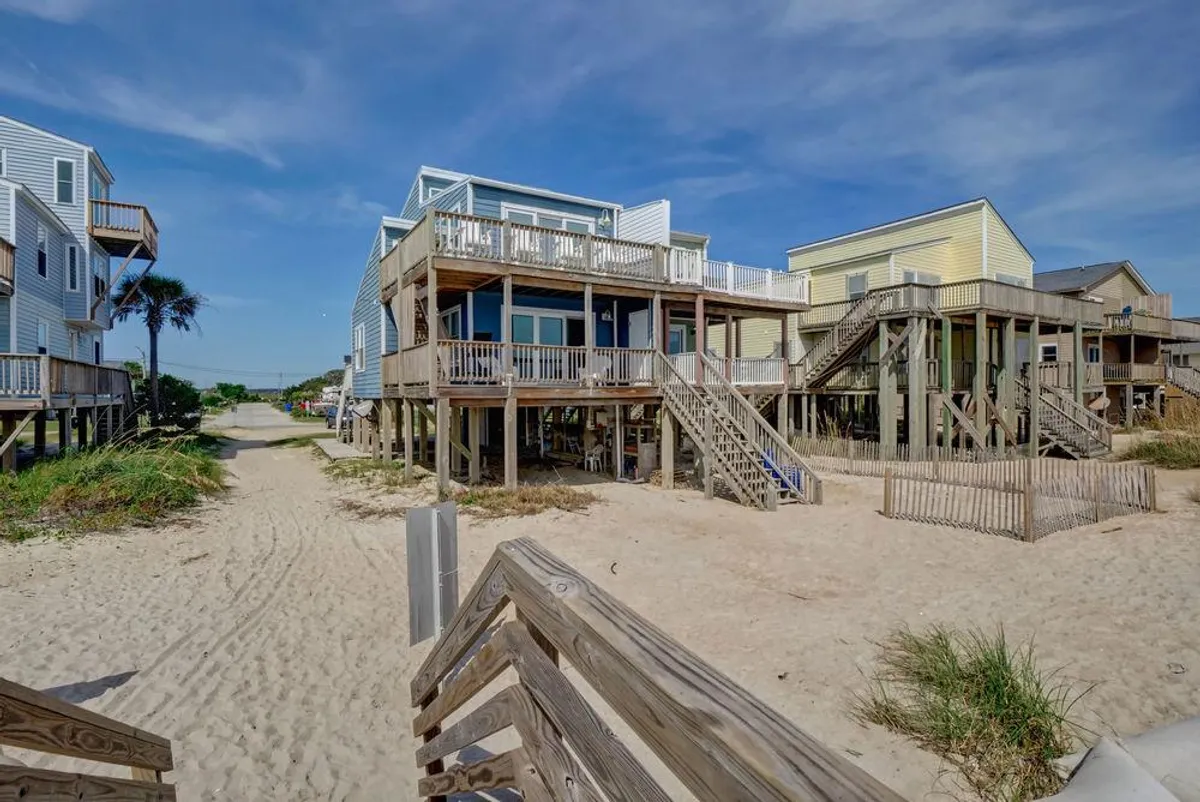 A view of classic beach houses on stilts with a sandy pathway leading directly to the expansive shoreline. Wooden walkways and porches evoke a sense of beachside living, with easy transitions between home and sand. The overall mood is casual and inviting, ideal for families stepping out for a day on the beach.