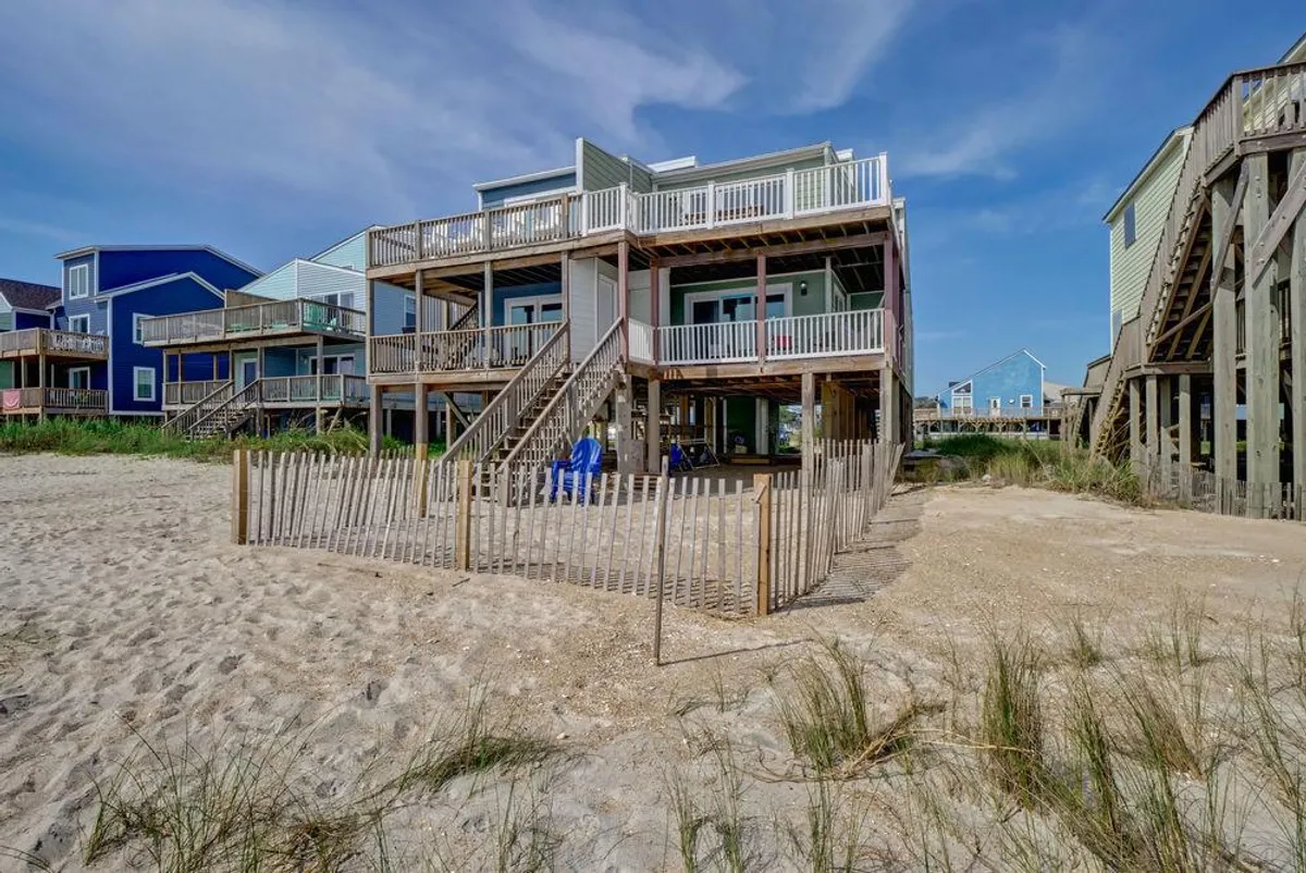 The image showcases a charming beachfront townhome with a fenced outdoor area, nestled among swaying beach grass and vibrant neighboring houses. The scene captures a laid-back atmosphere perfect for beach lounging, family gatherings, and soaking in the coastal ambiance.