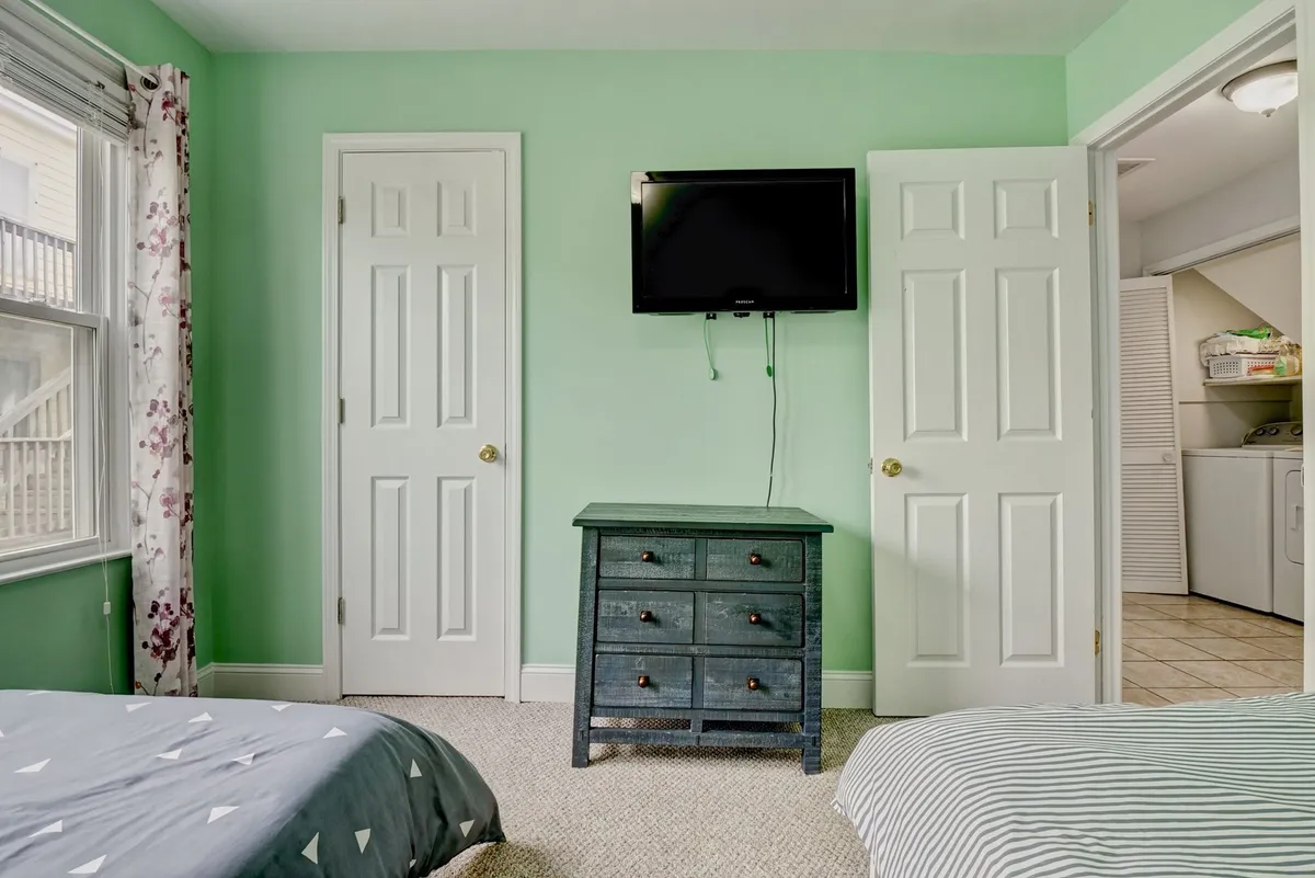 A cozy bedroom with green walls, floral curtains, a chest of drawers, and a TV, creating an inviting atmosphere for unwinding or late-night conversations.