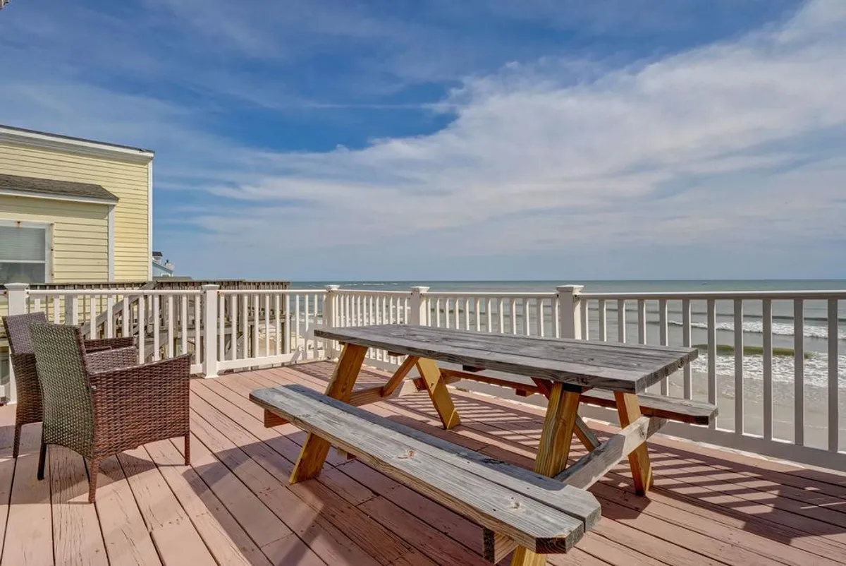 An inviting exterior setup with a wooden picnic table and wicker chairs overlooking a stunning ocean view. The space is perfect for outdoor dining, unwinding, or simply savoring the scenery as the sun sets over the water.