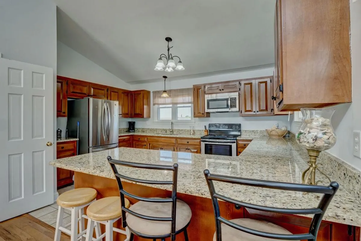 A bright, welcoming entryway and kitchen space featuring granite countertops, wooden cabinets, bar stools, and modern appliances. The layout suggests a seamless transition from entry to living areas, ideal for personalized routines.